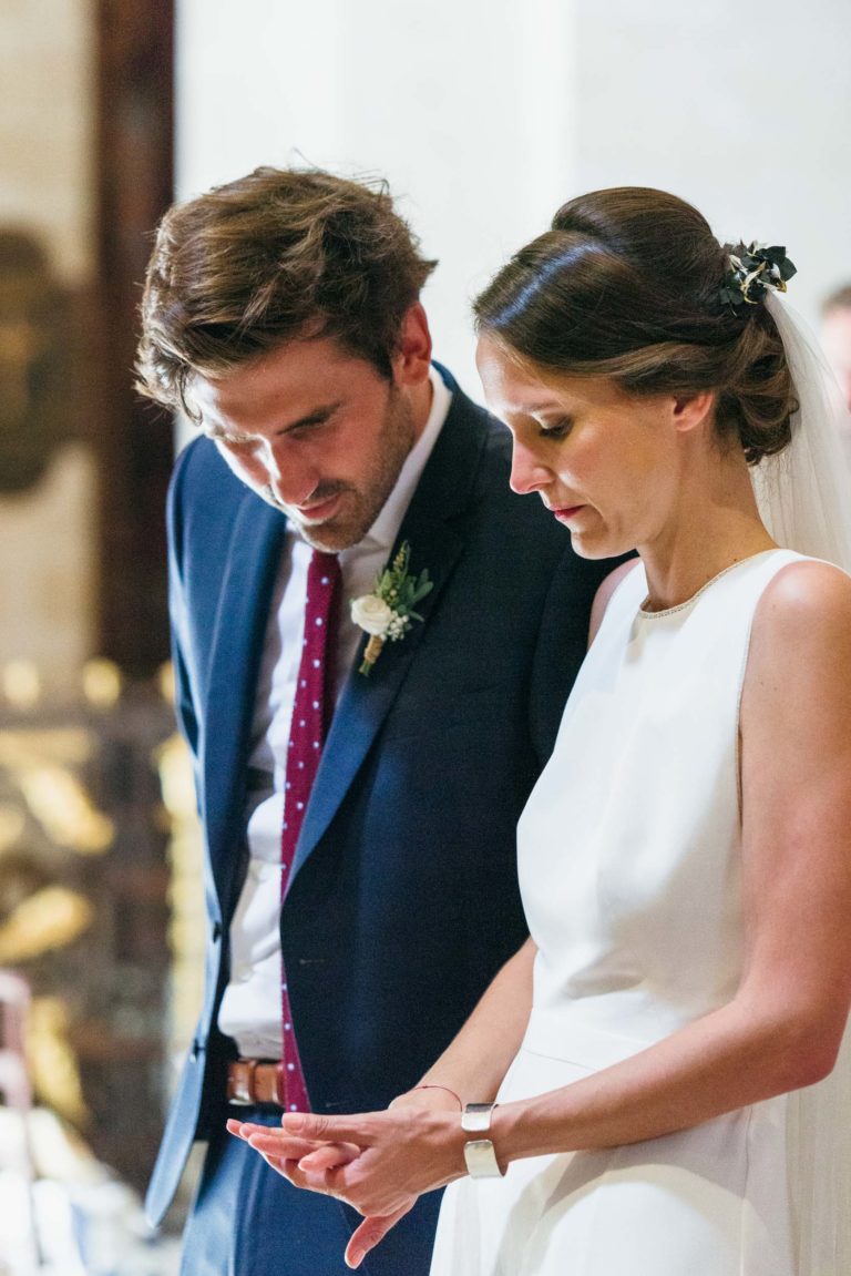 Un couple de mariés se tient côte à côte pendant leur mariage Parfait à Bordeaux. La mariée, vêtue d'une robe blanche avec un voile, regarde sa main. Le marié, vêtu d'un costume bleu foncé et d'une cravate rouge, regarde la main de la mariée. L'arrière-plan est légèrement flouté par un éclairage chaleureux et un mélange de tons sombres et clairs.