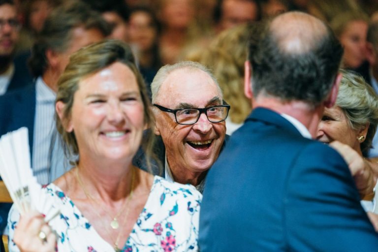 Un groupe de personnes est rassemblé étroitement dans une ambiance animée. Au premier plan, une femme souriante avec un haut fleuri tient plusieurs éventails en papier pliés. Au centre, un homme âgé portant des lunettes rayonne de joie. L'arrière-plan est rempli de personnes, suggérant un événement social comme le mariage Parfait à Bordeaux.