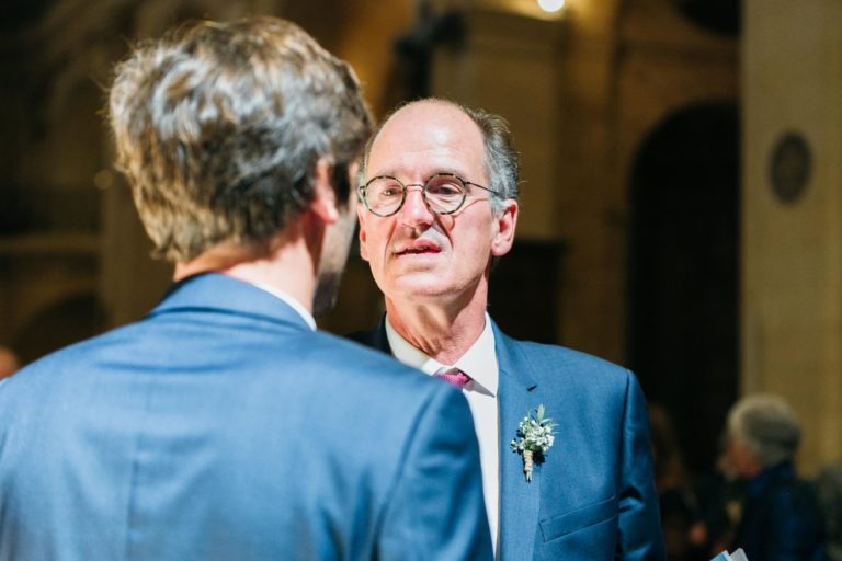 Deux hommes en costume discutent lors d'un événement en salle, dans le cadre du Mariage Parfait à Bordeaux. Face à la caméra, un homme avec des lunettes semble parler, vêtu d'un costume sombre, d'une chemise blanche, d'une cravate rose et d'une boutonnière fleurie. L'autre homme tourne le dos à la caméra. L'arrière-plan est faiblement éclairé, avec des figures floues et des détails architecturaux.