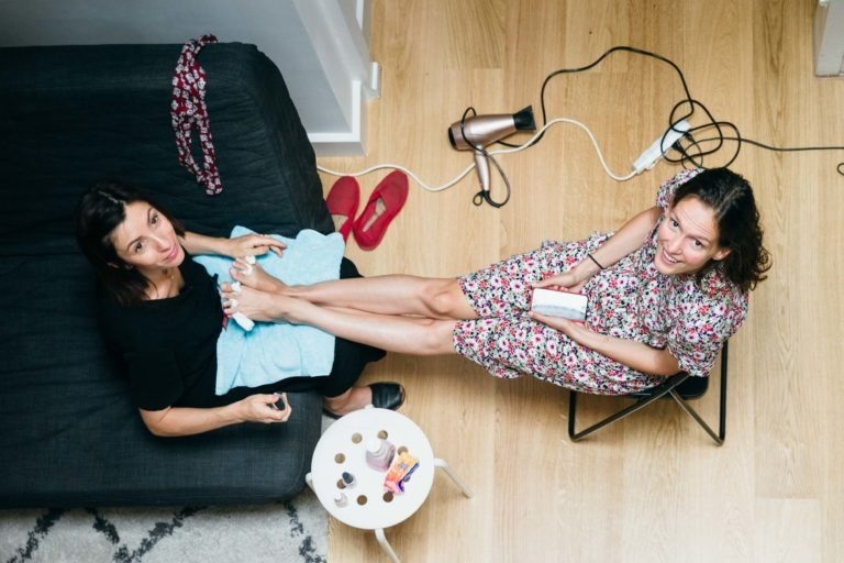 Deux femmes se font faire une pédicure assises dans un salon au parquet en bois. L'une, habillée de noir, applique du vernis à ongles à l'autre, qui porte une robe fleurie et est assise sur une petite chaise. Elles sourient à la caméra, évoquant la chaleur d'un mariage intime Parfait à Bordeaux.