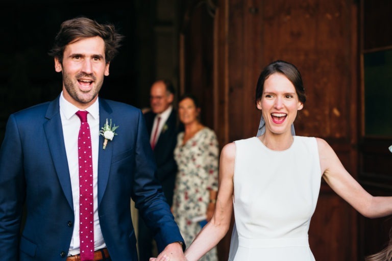 Un couple joyeux, main dans la main et sourire aux lèvres, sort d’un bâtiment aux portes en bois. L’homme, vêtu d’un costume bleu marine, d’une chemise blanche et d’une cravate à pois rouges, porte une boutonnière au revers de sa veste. La femme porte une robe blanche sans manches. C’est le mariage Parfait à Bordeaux. Deux silhouettes floues se tiennent à l’arrière-plan, habillées de façon formelle.