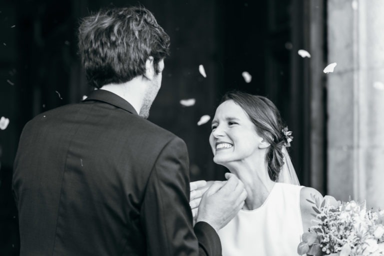 Photographie en noir et blanc d'un mariage parfait à Bordeaux, où la mariée, tenant un bouquet et souriant joyeusement, regarde le marié dos à la caméra. Des pétales sont jetés dans les airs autour d'eux, ajoutant à l'atmosphère de fête. La mariée porte un voile et le marié est en costume.