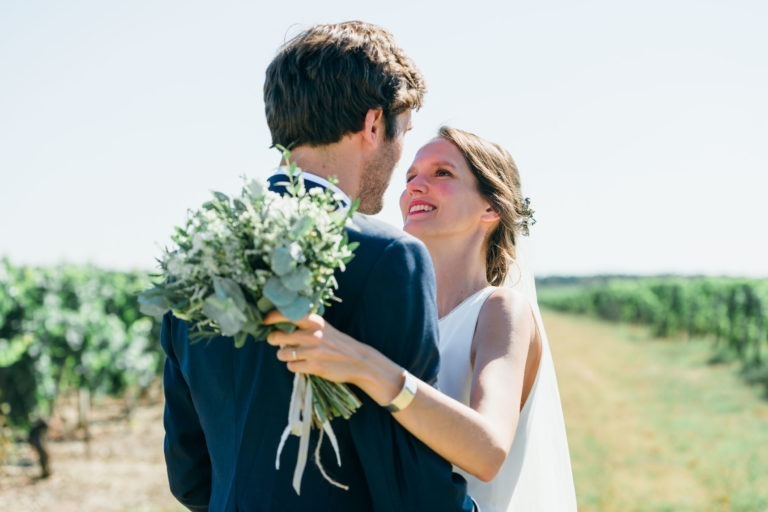 Un couple de mariés se tient face à face dans un vignoble, incarnant le mariage parfait à Bordeaux. La mariée, souriante avec un rouge à lèvres rouge vif, tient un bouquet de fleurs et porte une robe blanche avec un voile. Le marié, en costume sombre, tourne le dos à la caméra. Des vignes vertes s'étendent sous un ciel clair.