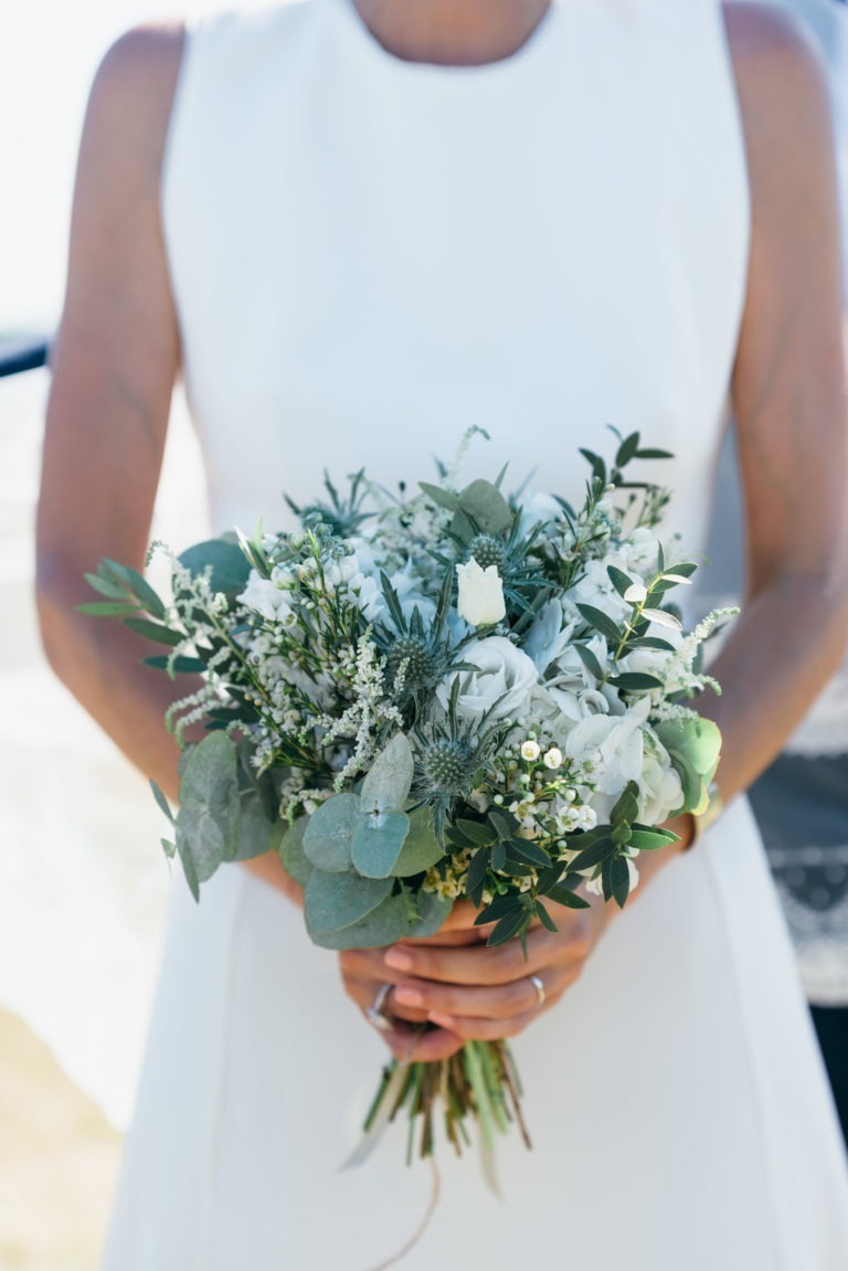 Une personne vêtue d'une robe blanche sans manches tient un bouquet de fleurs blanches et vertes. Le bouquet comprend un mélange de roses blanches, de feuilles d'eucalyptus et d'autres petits feuillages blancs et verts, créant un arrangement élégant et frais. L'arrière-plan flou suggère un décor extérieur, parfait pour un mariage Parfait à Bordeaux.