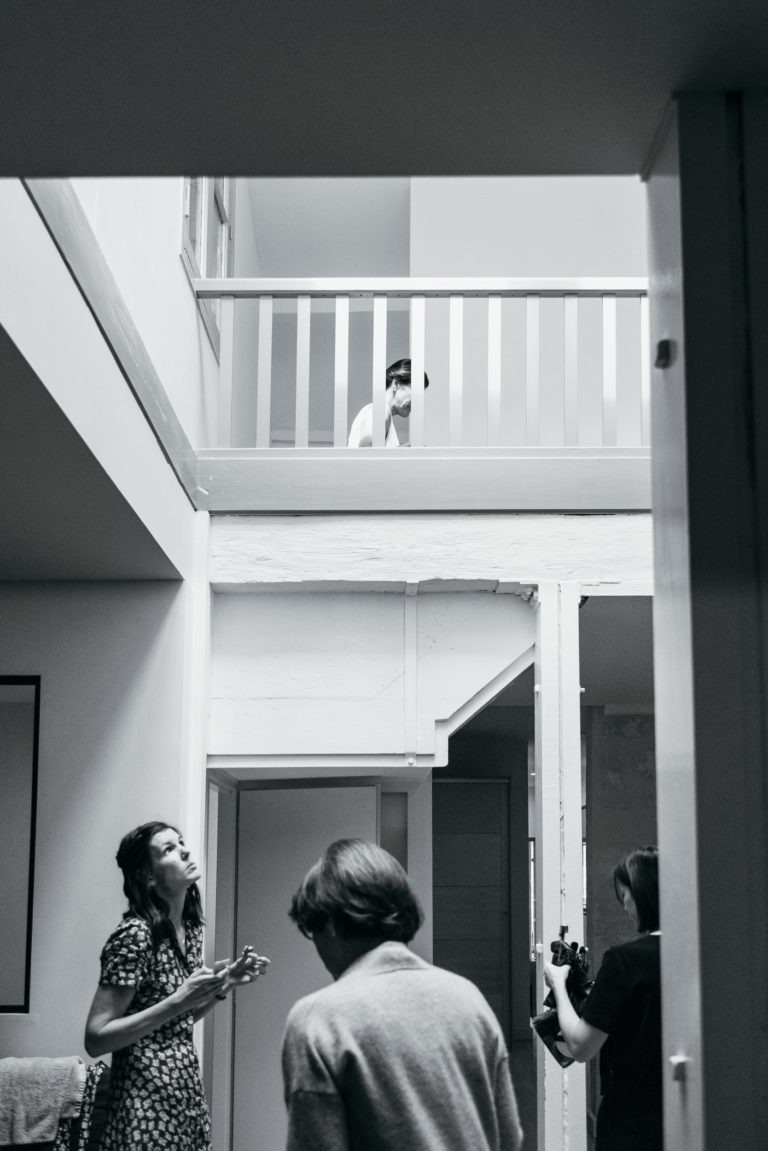 Une photo monochrome montre une scène d'intérieur avec trois personnes au rez-de-chaussée et une personne regardant vers le bas depuis un balcon à l'étage. Les individus au rez-de-chaussée sont engagés dans une conversation et semblent tenir des appareils photo ou d'autres équipements, capturant des moments d'un mariage parfait à Bordeaux. Le cadre est moderne et spacieux.