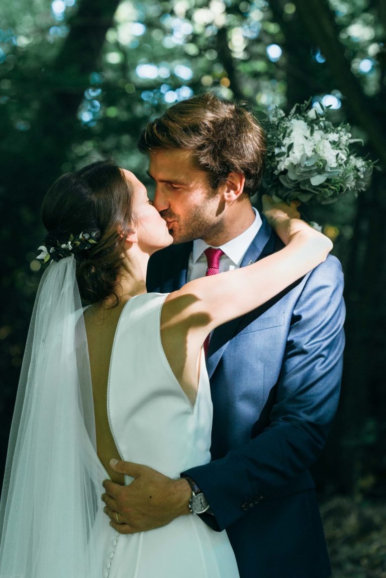 Un couple de mariés s'embrassent en plein air, incarnant l'essence même d'un mariage parfait à Bordeaux. La mariée porte une robe blanche à dos nu, ornée d'un voile décoré de petites fleurs. Le marié, en costume bleu et cravate rouge, la tient fermement tandis que la lumière du soleil filtre à travers les arbres derrière eux.