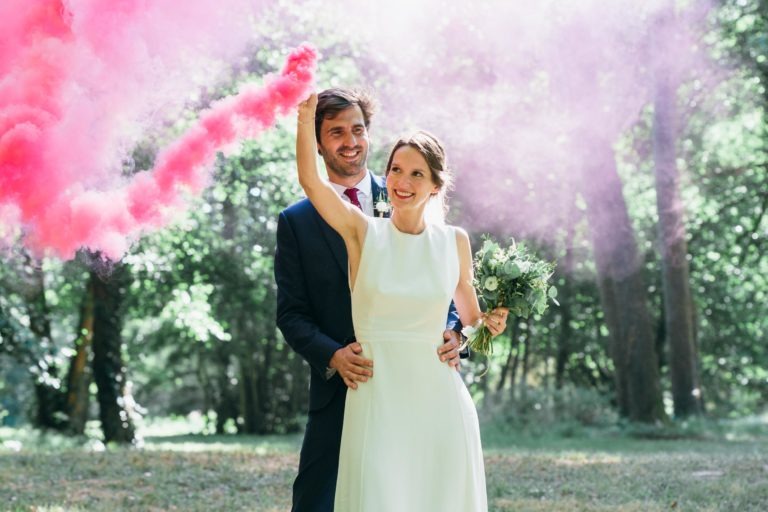 Un couple se tient dans une clairière, souriant. La femme, vêtue d'une robe blanche sans manches, tient une bombe fumigène émettant de la fumée rose. L'homme, vêtu d'un costume sombre, se tient derrière elle, les bras autour de sa taille. Le feuillage vert et la douce lumière du soleil créent un décor serein, un véritable mariage parfait à Bordeaux.