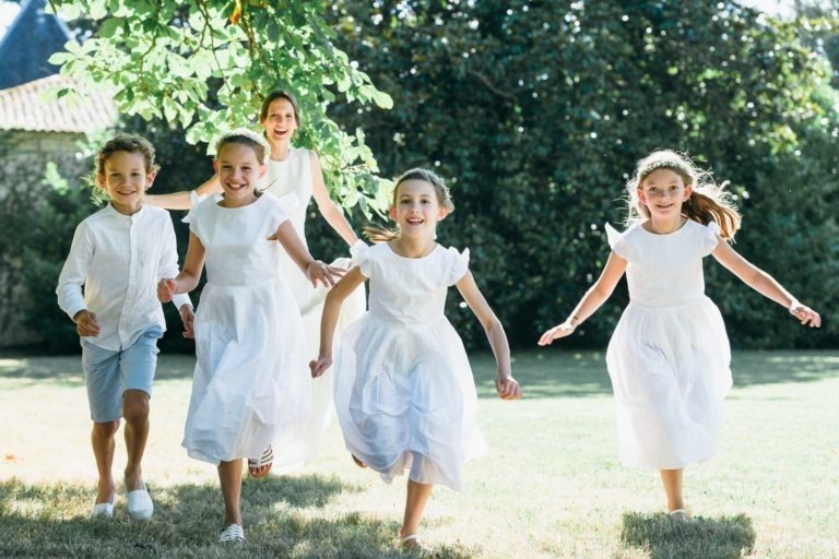 Un groupe joyeux d'enfants et une femme adulte courent vers la caméra dans un jardin herbeux et ensoleillé lors d'un mariage Parfait à Bordeaux. Les quatre enfants, trois filles en robe blanche et un garçon en chemise blanche et short bleu clair, ont des expressions heureuses. La femme à l'arrière-plan sourit et est habillée en blanc.