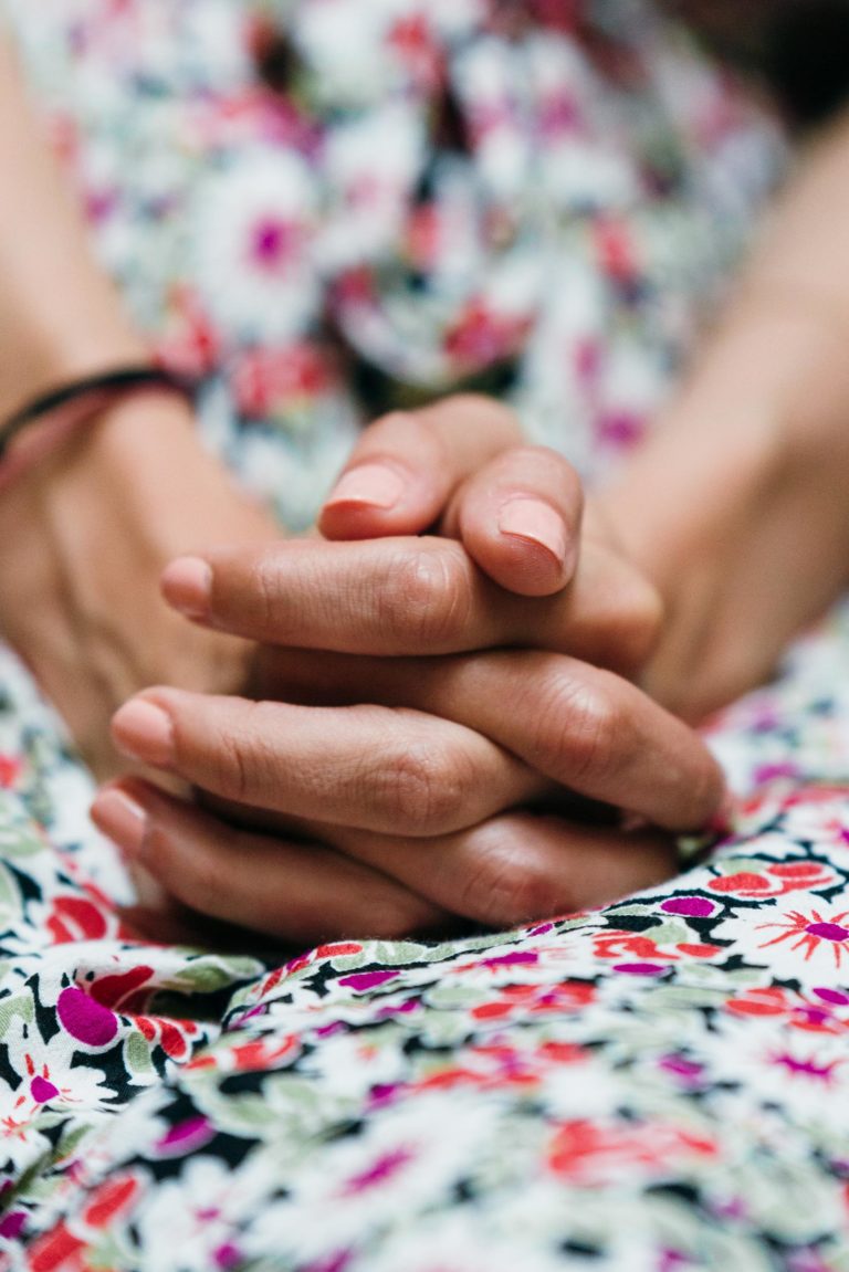 Gros plan sur les mains d'une personne posées sur ses genoux, les doigts entrelacés. Elle porte une robe fleurie au motif coloré composé de fleurs rouges, roses, blanches et vertes. L'arrière-plan est légèrement flou, mettant l'accent sur les mains et le tissu. La lumière est douce, ajoutant une ambiance douce, un véritable mariage parfait à Bordeaux.