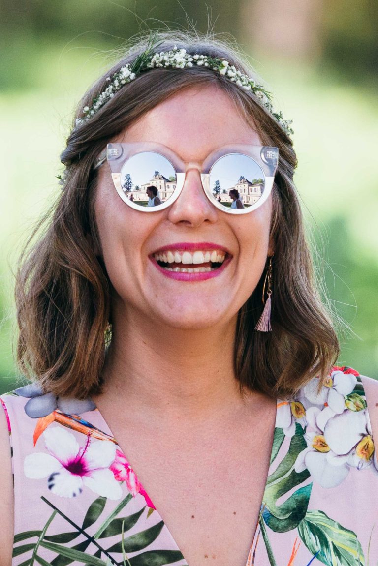 Une femme aux cheveux châtains mi-longs et coiffée d'un bandeau fleuri sourit largement. Elle porte des lunettes de soleil réfléchissantes qui laissent apparaître un bâtiment et de la verdure dans le reflet. Vêtue d'une tenue à motifs floraux pour le mariage Parfait à Bordeaux, elle porte une boucle d'oreille à pompon rose. L'arrière-plan est un paysage vert flou.