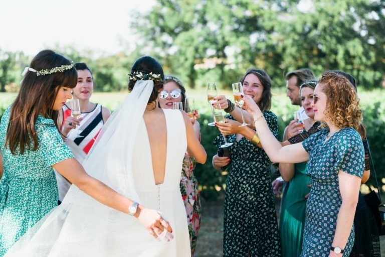 Une mariée en robe blanche et voile, vue de dos, trinque avec un groupe de femmes portant des robes colorées. Elles sont réunies à l'extérieur dans un cadre verdoyant et ensoleillé avec des arbres en arrière-plan. Les femmes tiennent des coupes de champagne et semblent joyeuses, célébrant ensemble leur mariage Parfait à Bordeaux.