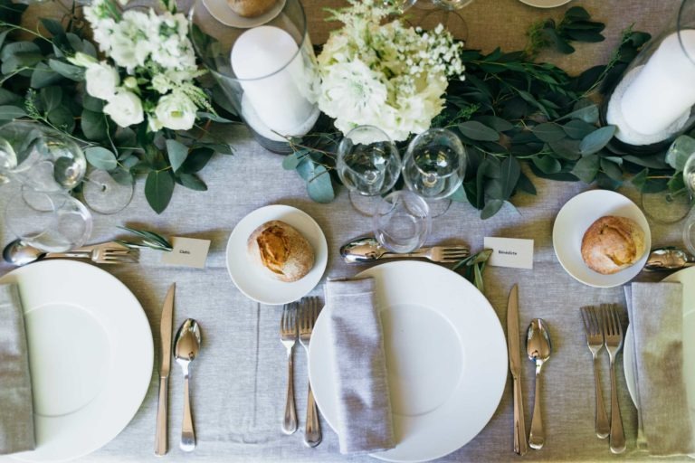 Une table joliment dressée pour un repas, avec des assiettes blanches, des couverts en argent et une serviette en tissu gris à chaque couvert. Chaque couvert est doté d'une carte de visite et d'un petit pain sur une petite assiette. La table, rappelant un parfait de mariage à Bordeaux, est ornée de verdure et de centres de table en fleurs blanches et de grandes bougies blanches dans des bougeoirs en verre.