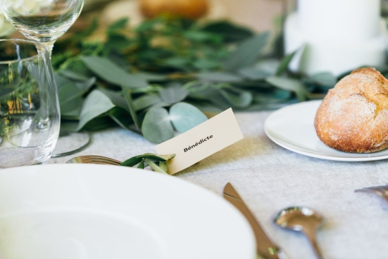 Gros plan sur une table dressée pour le mariage parfait à Bordeaux : une assiette blanche, un petit pain sur une petite assiette blanche, des couverts en argent et un verre à vin vide. Une carte avec le nom « Bénédicte » est posée sur la table, entourée de feuilles vertes utilisées comme décoration. La nappe est d'une couleur claire et neutre.