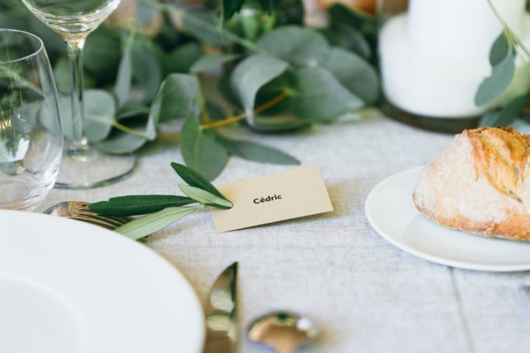 Une table dressée avec une assiette blanche, un couteau, une fourchette et un verre à vin. Une petite carte marron avec le nom « Cédric » écrit dessus est placée à côté d'un feuillage vert. Un petit pain rond est posé sur une assiette blanche à droite et la table est recouverte d'une nappe de couleur claire : un véritable parfait à Bordeaux pour un mariage.