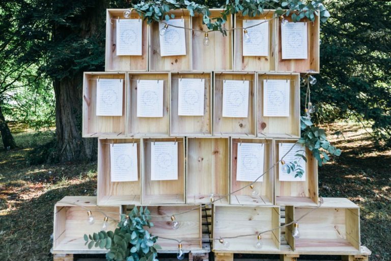 Des caisses en bois sont empilées pour créer un affichage à plusieurs niveaux avec des cartes imprimées suspendues à des ficelles devant chaque caisse. Les cartes semblent contenir du texte, probablement pour la disposition des sièges ou des messages lors d'un mariage Parfait à Bordeaux. Des feuilles et une guirlande lumineuse ornent l'affichage, sur fond d'arbres extérieurs.