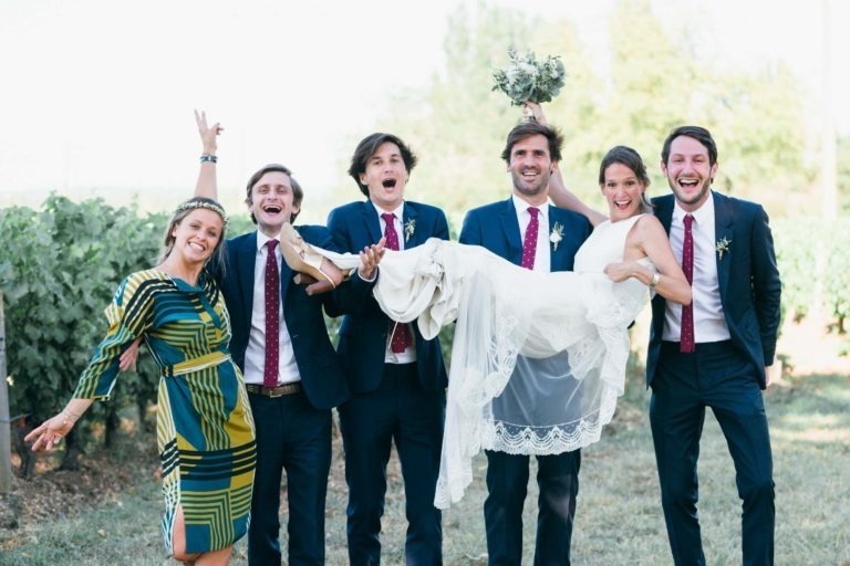 Une photo de groupe montre six adultes en plein air dans un vignoble. Quatre hommes en costume bleu marine et cravate rouge tiennent une femme en robe de mariée blanche à l'horizontale. Une autre femme en robe colorée se tient sur le côté, les bras levés. Tout le monde sourit et semble célébrer un mariage parfait à Bordeaux.