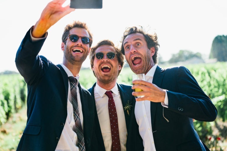 Trois hommes en costume et cravate prennent un selfie en plein air par une journée ensoleillée. Ils semblent excités, avec un large sourire et une expression joyeuse. L'un d'eux tient un smartphone pour le selfie, et l'autre tient un verre. Ils se tiennent devant un vignoble verdoyant et luxuriant, un mariage parfait à Bordeaux.