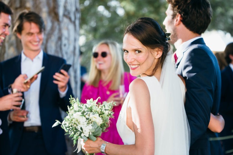 Une mariée en robe blanche et voile sourit à la caméra tout en tenant un bouquet de fleurs blanches, incarnant l'essence du mariage Parfait à Bordeaux. Elle est entourée d'invités au mariage : un homme en costume bleu marine tenant un téléphone, une femme en tenue rose vif portant des lunettes de soleil et un autre homme en costume bleu marine. Un tronc d'arbre est visible en arrière-plan.