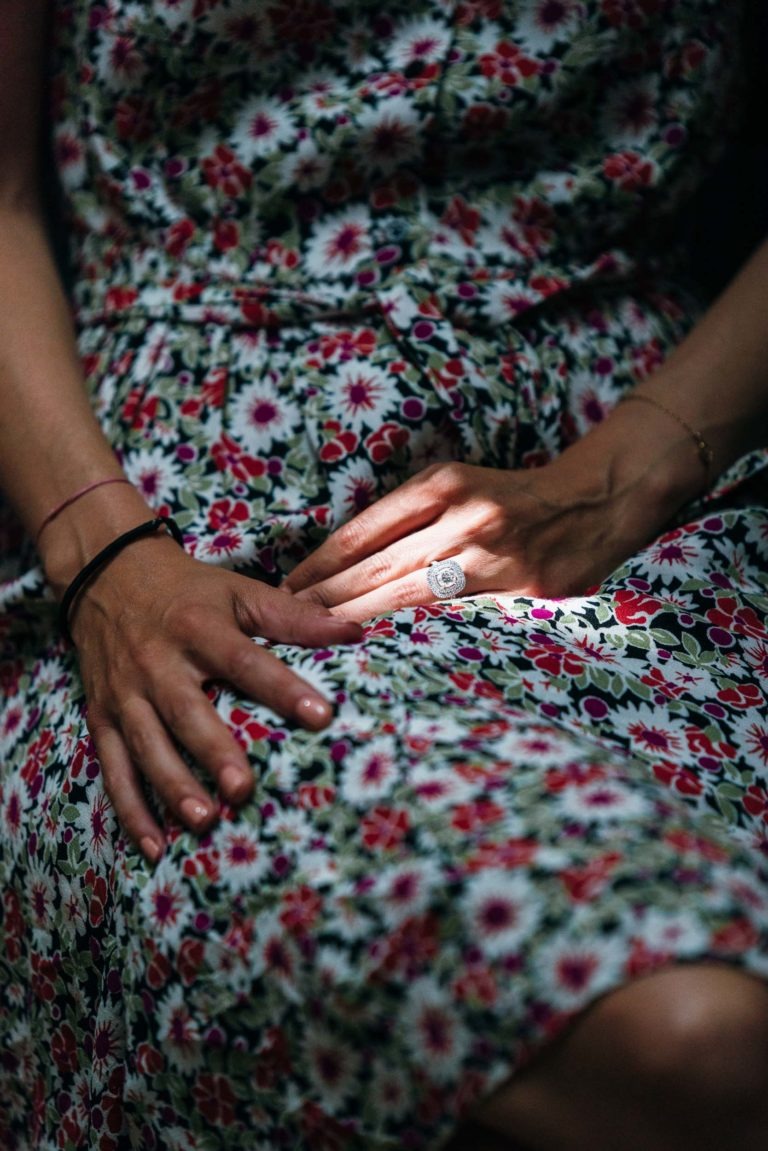 Gros plan d'une personne posant ses mains sur ses genoux, vêtue d'une robe fleurie ornée de petites fleurs roses, blanches et rouges et de feuilles vertes. Une bague proéminente sur son doigt capte la lumière. L'ambiance générale du mariage Parfait à Bordeaux est tamisée, avec un faisceau de lumière mettant en valeur la bague et les mains.