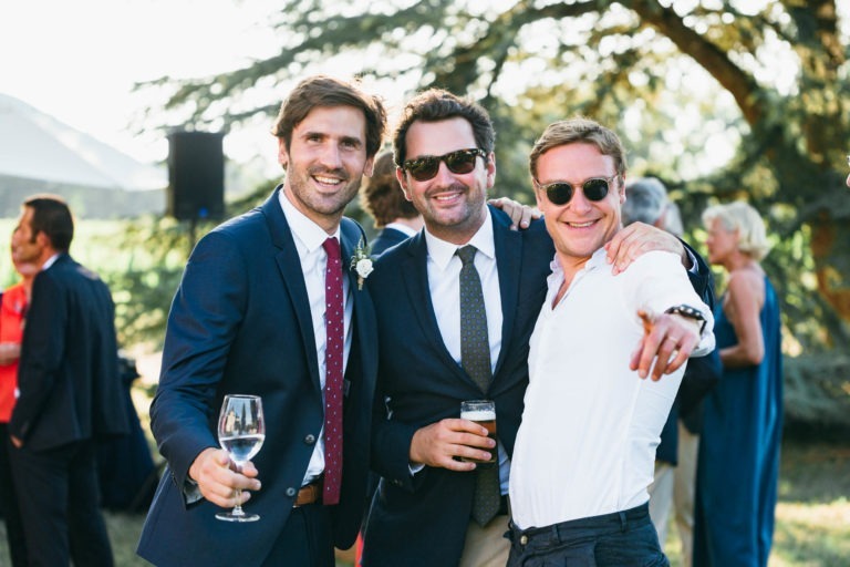 Trois hommes posent à l'extérieur dans un environnement festif, peut-être lors d'un mariage Parfait à Bordeaux. Ils sont habillés de manière semi-formelle, deux d'entre eux en costume et le troisième en chemise blanche. Chacun tient un verre, souriant et s'amusant. L'arrière-plan présente des arbres et d'autres personnes en conversation.