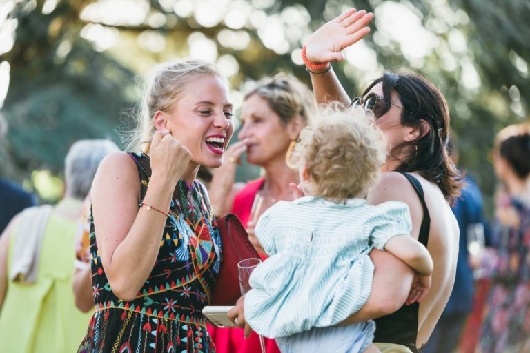 Un rassemblement extérieur animé avec quatre femmes, l'une tenant un jeune enfant aux cheveux bouclés. Les femmes sont engagées dans une conversation animée et des rires, rappelant un mariage parfait à Bordeaux. Une femme porte une robe colorée et sourit largement tandis qu'une autre a le bras levé, indiquant l'excitation ou l'emphase. De la verdure est visible en arrière-plan.