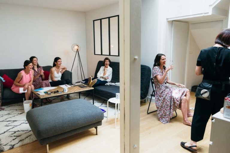 Un groupe de six femmes se trouve dans un salon. Quatre d'entre elles sont assises sur un canapé et une autre sur le sol rient ensemble, profitant d'une atmosphère détendue rappelant le Mariage Parfait à Bordeaux. Elles sont habillées de façon décontractée dans cette pièce décorée simplement, avec un lampadaire, un pouf gris et un miroir mural à compartiments. Une autre femme discute avec elles.