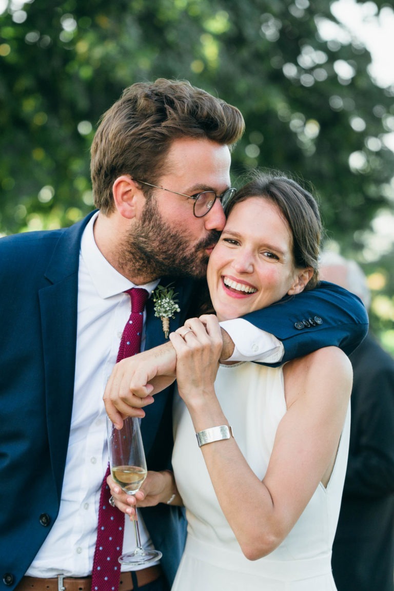 Un homme portant des lunettes et une barbe, vêtu d'un costume bleu et d'une cravate rouge, embrasse la joue d'une femme souriante portant une robe blanche sans manches. La femme tient un verre de champagne et se penche dans l'étreinte. Ils sont à l'extérieur avec de la verdure en arrière-plan, posant joyeusement pour leur Mariage Parfait à Bordeaux.