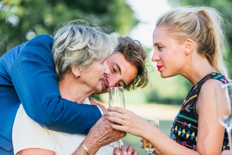 Un jeune homme en costume bleu serre dans ses bras une femme âgée aux cheveux gris par derrière, qui tient un verre de champagne. À côté d'eux, une jeune femme aux cheveux blonds coiffés en chignon haut tient un autre verre de champagne. Ils célèbrent ce qui semble être le mariage Parfait à Bordeaux en plein air, par une journée ensoleillée, avec un arrière-plan vert et flou.