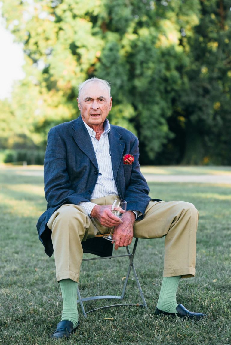 Un homme âgé aux cheveux gris courts est assis sur une chaise pliante à l'extérieur. Il porte un blazer bleu marine, une chemise bleu clair, un pantalon beige, des chaussettes vert clair et des chaussures à enfiler noires. Une de ses mains tient un verre à vin du mariage Parfait à Bordeaux. L'arrière-plan présente des arbres et de l'herbe verts luxuriants, suggérant un décor de parc.