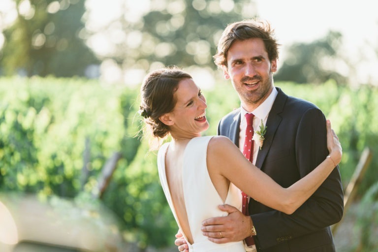 Une mariée en robe blanche sans manches et un marié en costume sombre avec une cravate à pois rouges s'embrassent joyeusement dans un vignoble. La mariée rit et regarde par-dessus son épaule, tandis que le marié sourit chaleureusement. Les vignes ensoleillées et l'arrière-plan flou créent une atmosphère sereine et romantique, un véritable mariage parfait à Bordeaux.