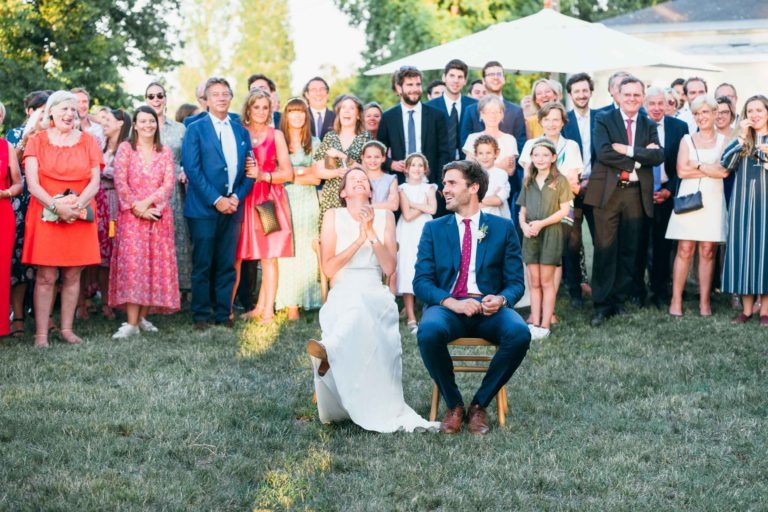 Un couple de mariés, tous deux souriants, sont assis sur des chaises en bois dans un espace extérieur gazonné pendant leur mariage parfait à Bordeaux. La mariée porte une robe blanche et le marié porte un costume bleu avec une cravate rouge. Derrière eux se tient un grand groupe d'invités en tenue colorée, tous regardant et souriant. Des arbres et une voûte blanche sont visibles en arrière-plan.