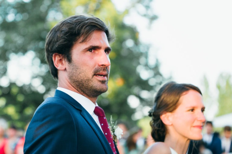 Un homme en costume bleu avec une cravate rouge et une femme avec une coiffure tressée sont vus à l'extérieur lors de ce qui semble être le Mariage Parfait à Bordeaux. L'homme regarde légèrement vers la gauche avec une expression sérieuse, tandis que la femme, légèrement devant lui, sourit doucement. L'arrière-plan est flouté par la verdure et une lumière naturelle vive.