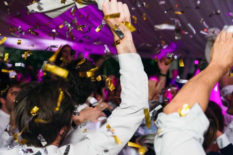 Les gens sont vus en train de faire la fête sous un dais de fête au Mariage Parfait à Bordeaux. Des confettis dorés, argentés et blancs tombent en cascade tandis que les gens lèvent les mains avec enthousiasme. La scène est festive, avec un éclairage violet vibrant en arrière-plan qui ajoute à l'atmosphère animée.