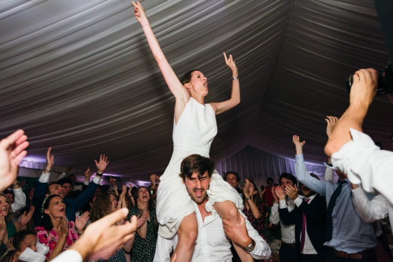 Une femme en robe blanche est portée sur les épaules d'un homme sous une grande tente blanche au Mariage Parfait à Bordeaux. Elle jubilait, un bras levé et la bouche ouverte. L'homme sourit et la foule autour d'elle applaudit et acclame, créant une atmosphère animée et festive.