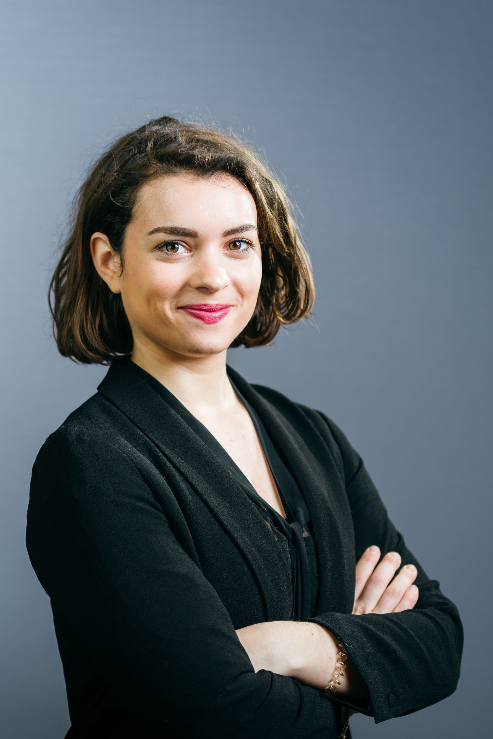 Une femme aux cheveux châtains mi-longs, vêtue d'un blazer noir et d'un haut foncé, se tient debout, les bras croisés. Elle sourit avec assurance sur un fond gris uni dans ce portrait d'entreprise. Elle porte un maquillage léger et du rouge à lèvres rose, ce qui reflète son professionnalisme.