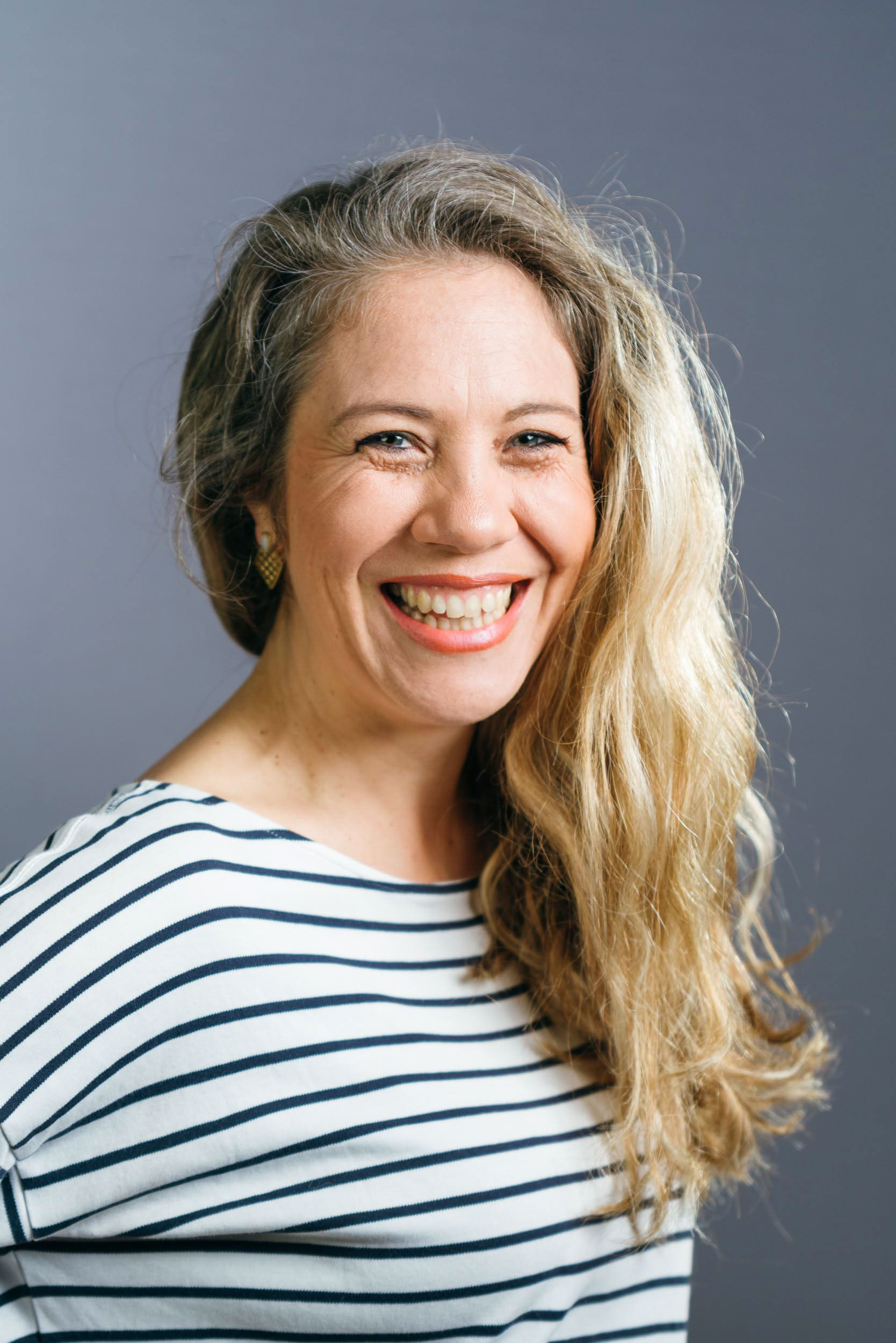 Dans ce portrait professionnel, une femme aux cheveux blonds longs et ondulés sourit largement à l'objectif. Elle porte un haut rayé blanc et bleu marine et se tient debout sur un fond gris uni, rayonnant d'une expression joyeuse et amicale - un portrait parfait pour les profils d'entreprise à Bordeaux, en Gironde.