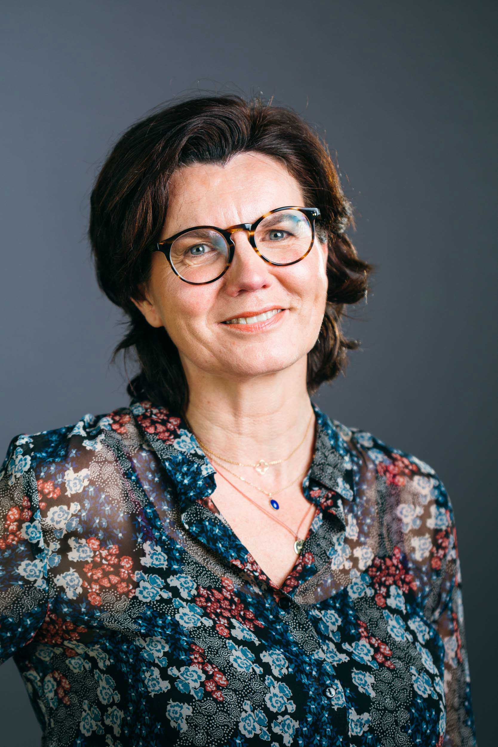 Une femme souriante aux cheveux courts et foncés et portant des lunettes est représentée sur un fond gris. Elle porte un chemisier fleuri semi-transparent avec un col à volants et un collier en or avec un pendentif. Son expression est amicale et accessible, parfaite pour un portrait d'entreprise dans le milieu professionnel de Bordeaux.