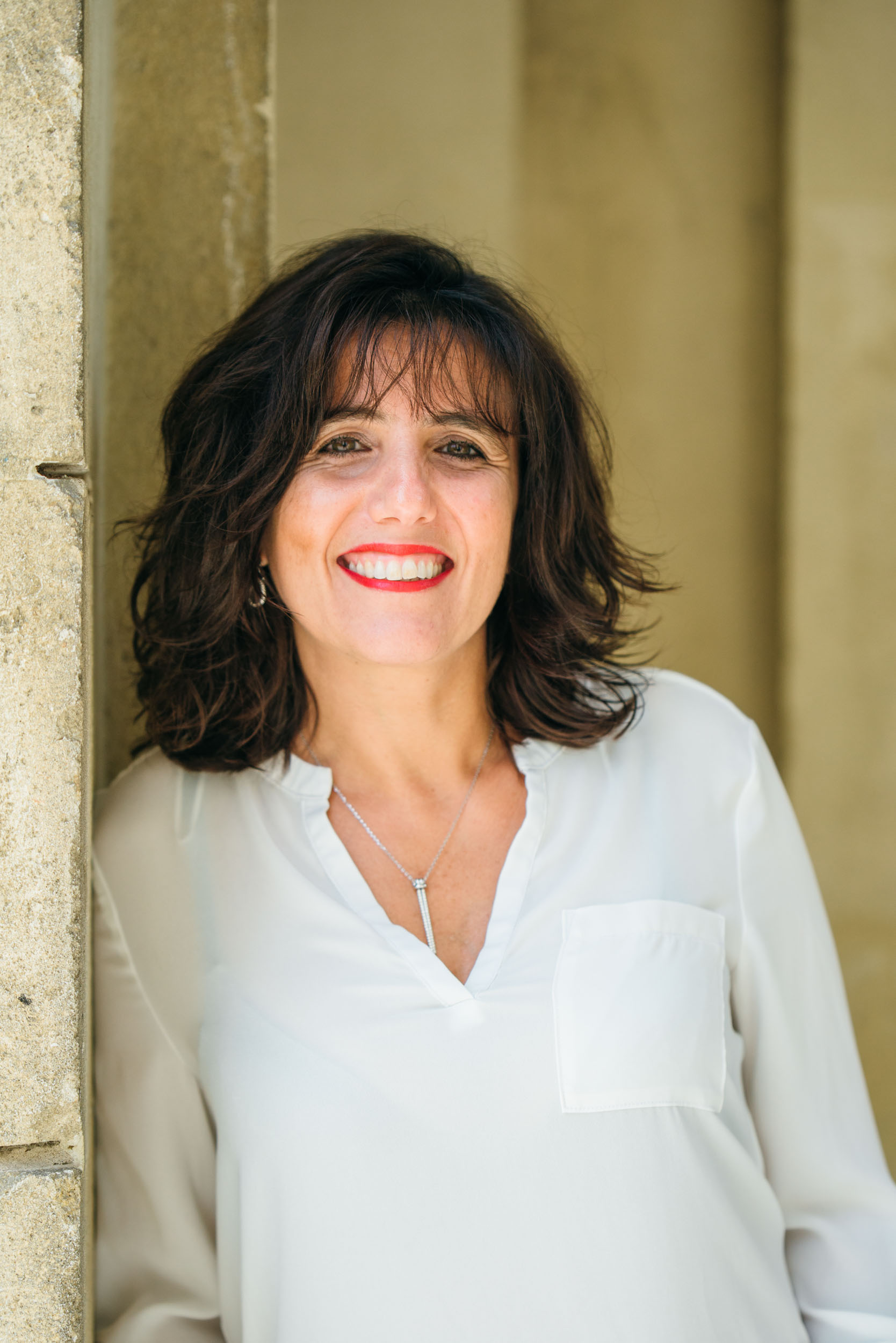 Une femme aux cheveux noirs mi-longs, vêtue d'un chemisier blanc à col en V et à poche avant, se tient debout, souriante, devant un mur en pierre beige à Bordeaux, en Gironde. Elle porte du rouge à lèvres rouge et un collier argenté qui complète sa tenue. Le décor général semble être extérieur avec un éclairage naturel, parfait pour un portrait d'entreprise.