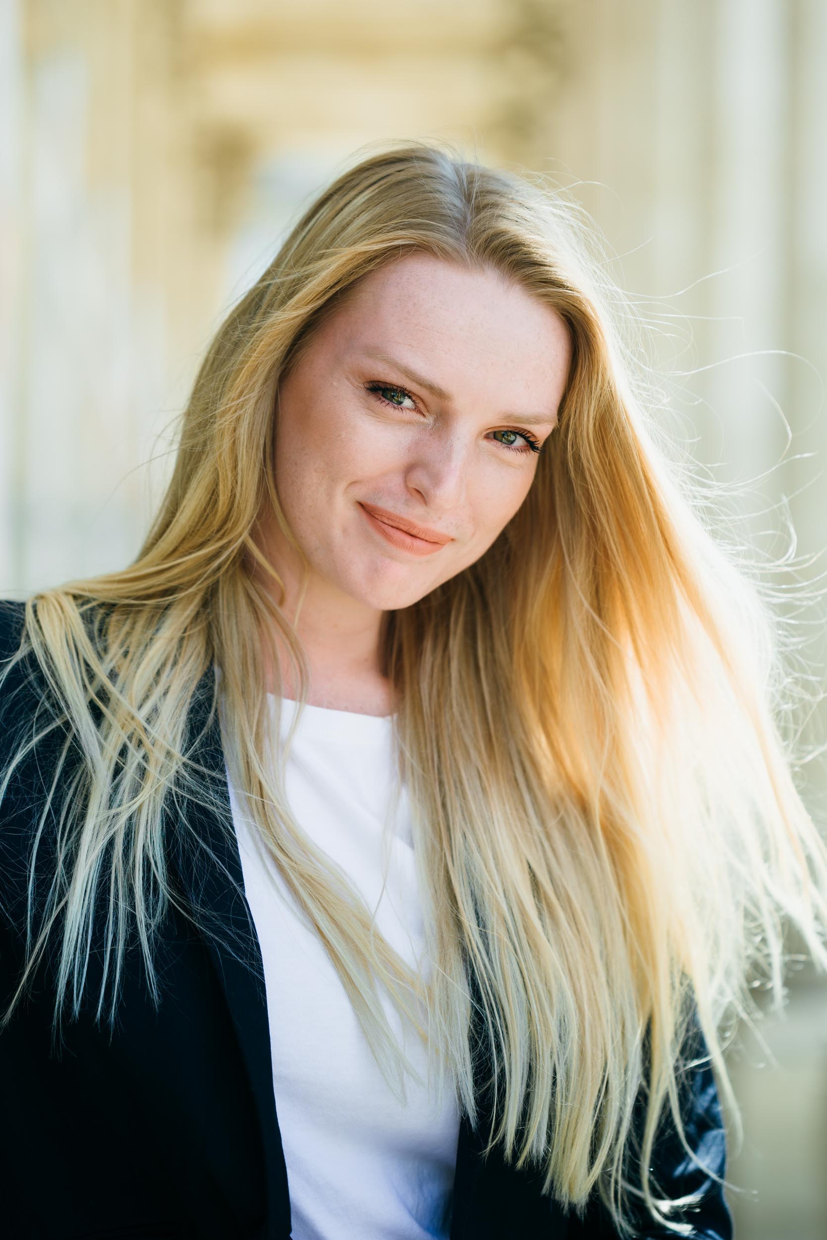 Une femme aux cheveux longs et blonds sourit légèrement en regardant l'objectif. Elle porte un blazer noir sur un haut blanc. L'arrière-plan est flou, avec un couloir ensoleillé ou une allée extérieure à Bordeaux, offrant une atmosphère aérée et lumineuse parfaite pour un portrait professionnel d'entreprise.