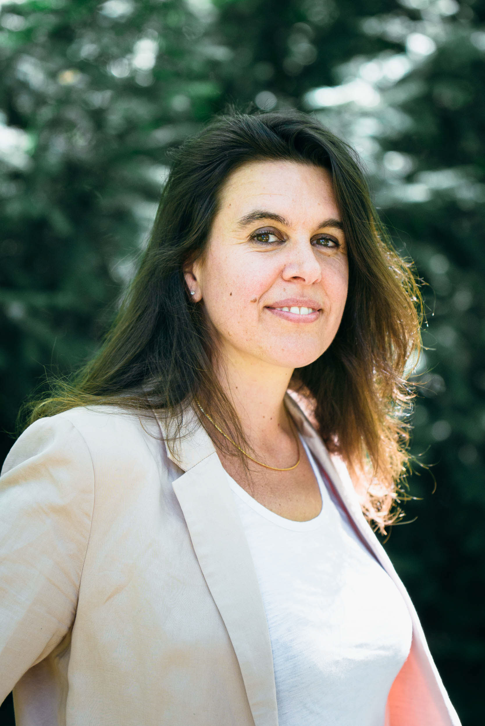 Une femme aux longs cheveux bruns se tient debout à l'extérieur, souriant légèrement. Vêtue d'un blazer beige clair sur une chemise blanche, elle dégage une élégance professionnelle. La verdure floue à l'arrière-plan suggère un jardin ou une forêt. Ce portrait professionnel a été pris avec un éclairage naturel et lumineux qui illumine son visage et ses cheveux.