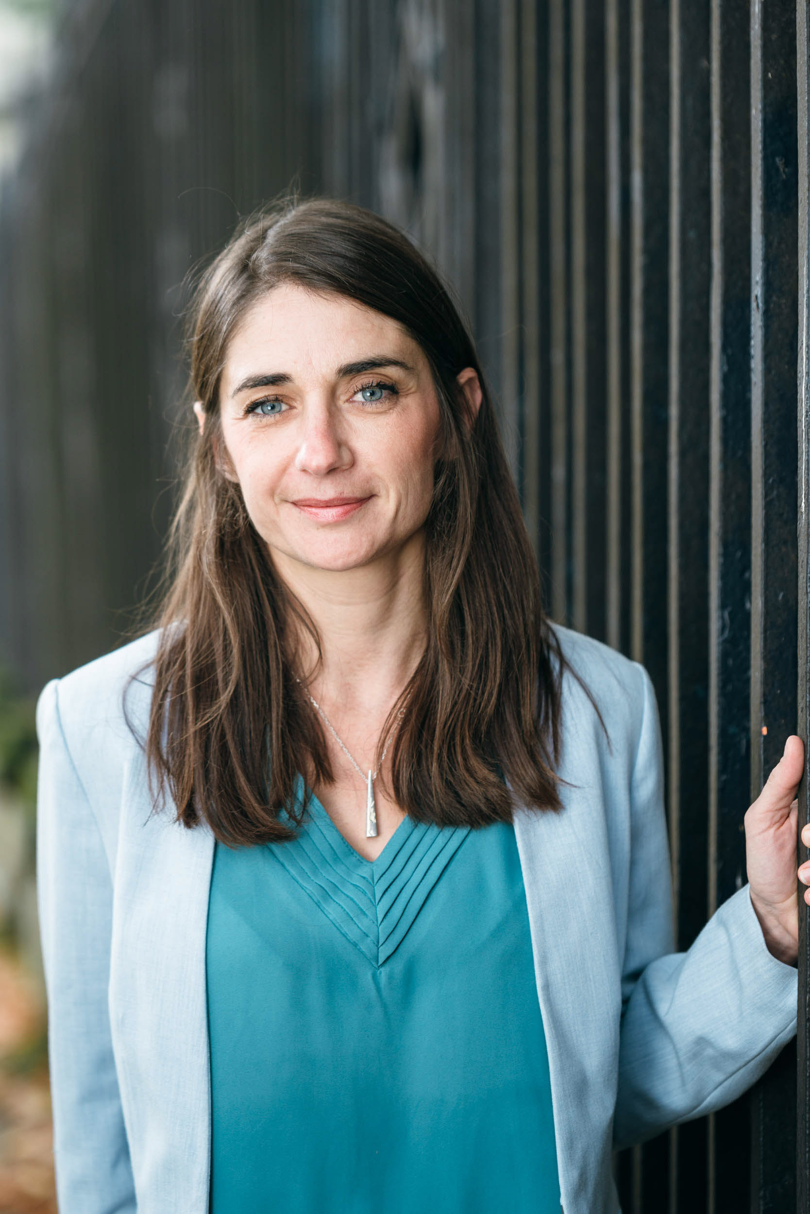 Une femme aux longs cheveux bruns et aux yeux bleus se tient debout près d'une clôture en métal noir, regardant la caméra avec un doux sourire. Dans ce portrait d'entreprise, elle porte un blazer bleu clair sur un chemisier bleu canard, complété par un collier en argent. Le décor de Bordeaux, en Gironde, ajoute une touche d'élégance à la scène.