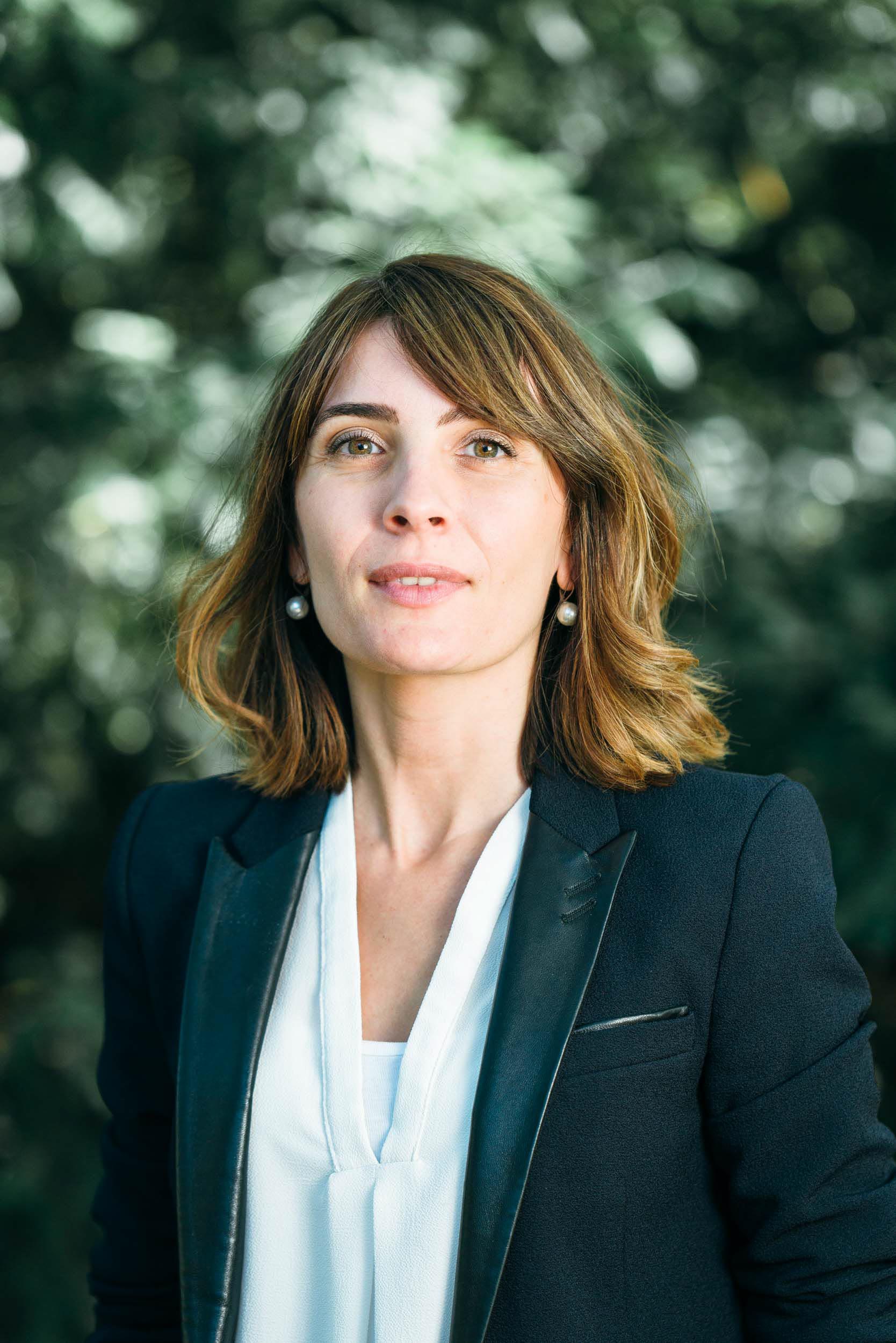 Une femme aux cheveux châtains mi-longs, vêtue d'un blazer noir et d'un chemisier blanc, se tient debout à l'extérieur, devant un arrière-plan flou de feuillage vert. Elle a une expression neutre et porte des boucles d'oreilles en perles, regardant directement l'appareil photo dans ce portrait d'entreprise professionnel pris à Bordeaux, en Gironde.