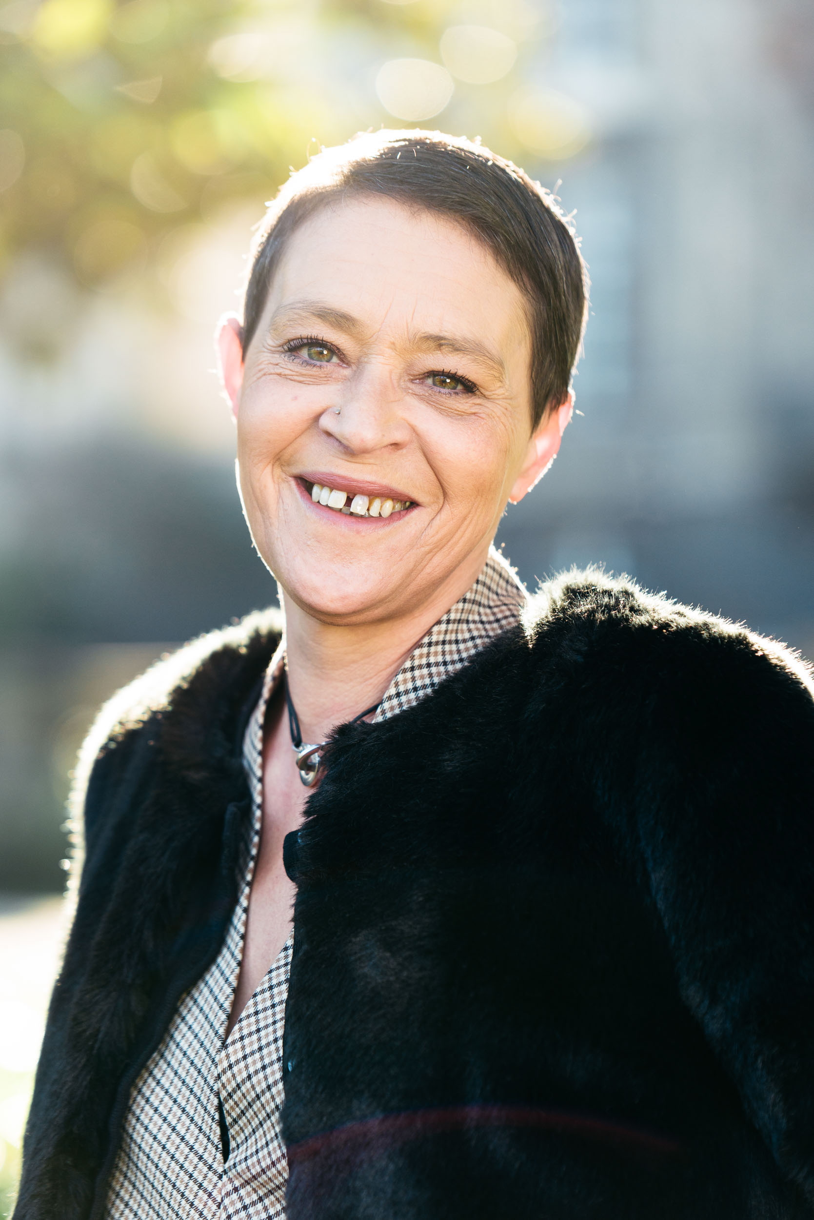 Une femme aux cheveux noirs courts sourit à l'objectif. Vêtue d'une chemise à carreaux et d'une veste en peluche sombre, elle respire la confiance dans ce portrait d'entreprise. L'arrière-plan légèrement flou laisse entrevoir la verdure luxuriante et la lumière chaude du soleil de Bordeaux, en Gironde, créant une ambiance radieuse.