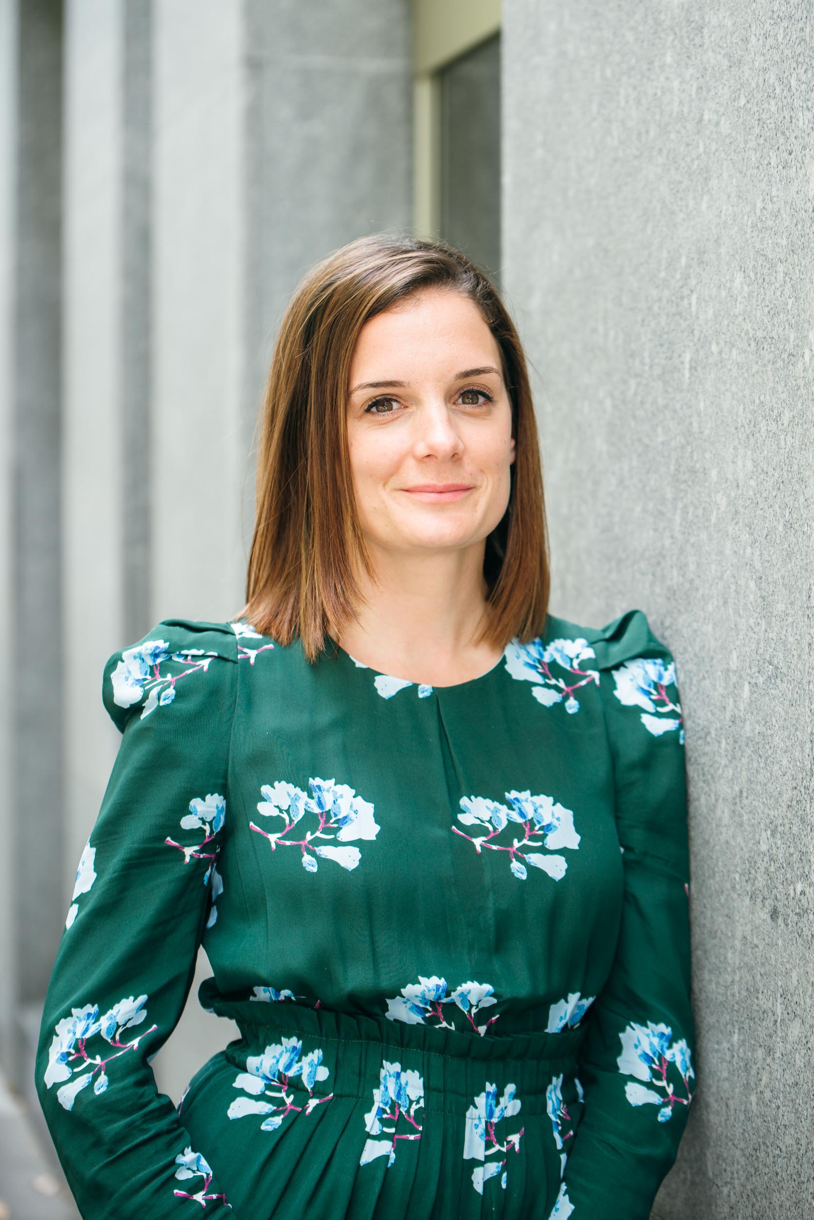 Une femme aux cheveux bruns raides et mi-longs se tient debout contre un mur gris à Bordeaux, souriant doucement. Elle porte une robe vert foncé avec des imprimés floraux bleus et blancs et a un maquillage subtil. L'arrière-plan est légèrement flou, se concentrant sur elle dans ce portrait professionnel comme sujet principal.