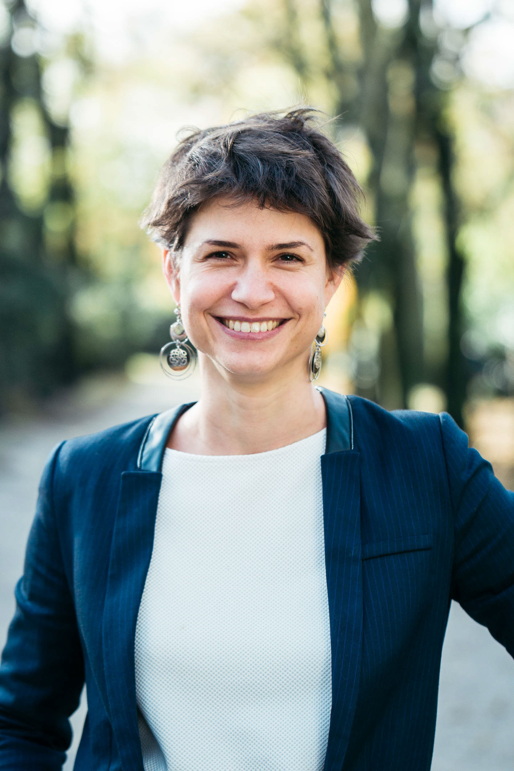 Une personne aux cheveux bruns courts sourit à l'objectif sur ce portrait d'entreprise. Elle porte un blazer bleu marine sur un haut blanc et des boucles d'oreilles pendantes en argent. Sur fond de feuillage vert et d'arbres, elle dégage une attitude amicale et accessible, parfaite pour tout environnement professionnel à Bordeaux Gironde.