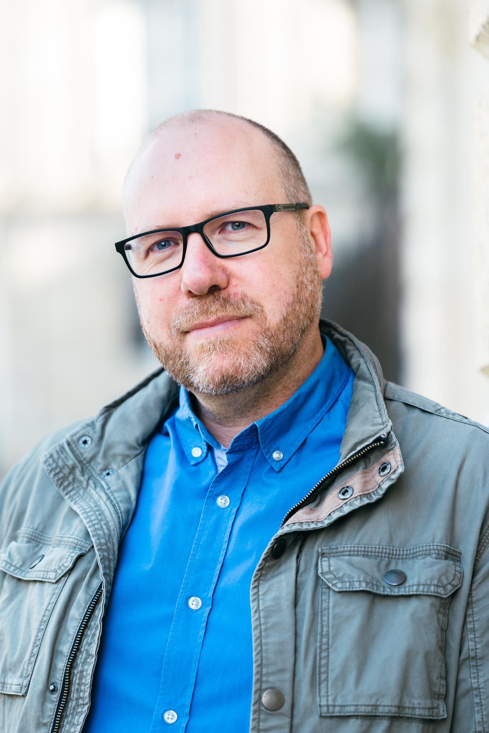 Un homme d'âge moyen au crâne chauve, à la barbe claire et aux lunettes, porte une chemise bleue sous une veste grise. Il se tient à l'extérieur dans une lumière douce et naturelle avec une expression neutre. Ce portrait d'entreprise a été pris à Bordeaux, en Gironde, avec une structure ou un bâtiment flou en arrière-plan.