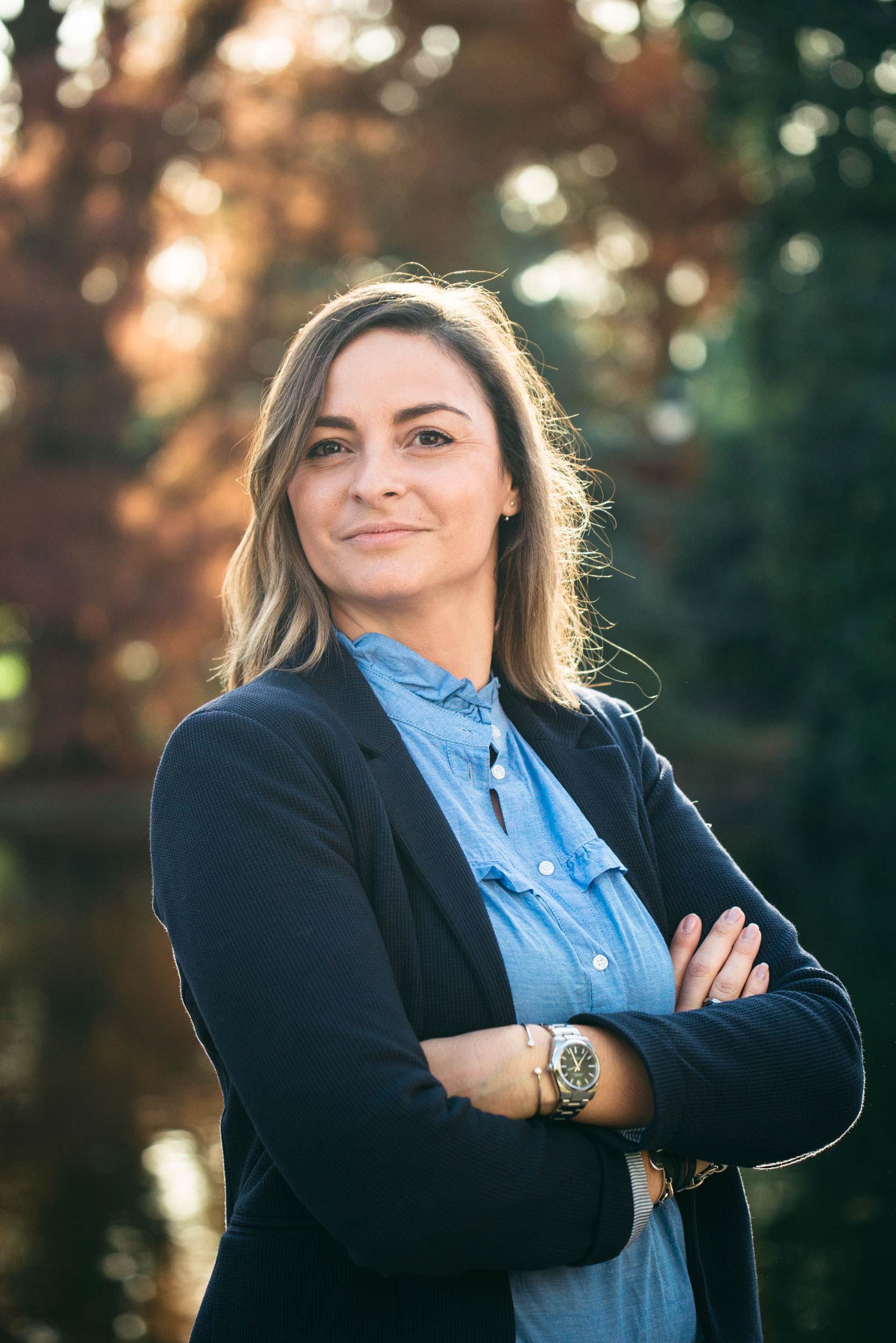 Une femme confiante aux cheveux bruns mi-longs se tient debout, les bras croisés, dans ce portrait professionnel. Vêtue d'une chemise bleue boutonnée et d'un blazer noir, elle est à l'extérieur, avec des arbres flous et la lumière du soleil en arrière-plan, créant une atmosphère sereine rappelant Bordeaux.