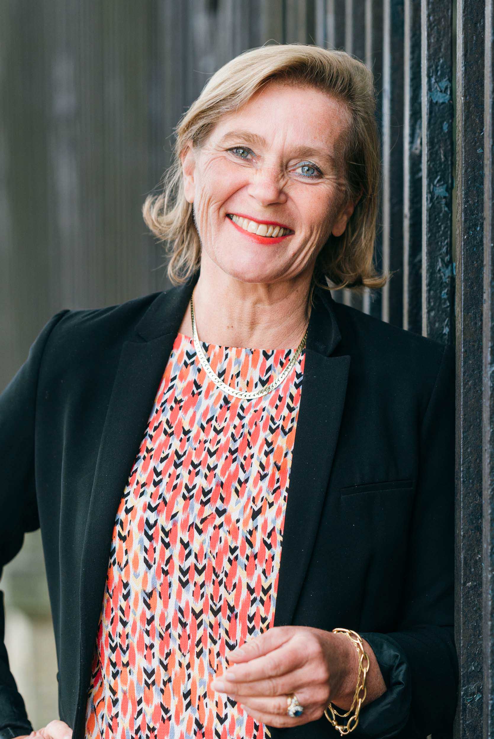 Une femme souriante aux cheveux blonds courts s'appuie contre une structure sombre et verticale. Vêtue d'un blazer noir sur un haut coloré à motifs aux nuances de rose, de blanc et de noir, elle accessoirise d'un collier et d'un bracelet en or tout en tenant ses lunettes dans sa main droite, une parfaite portraitiste professionnelle à Bordeaux.