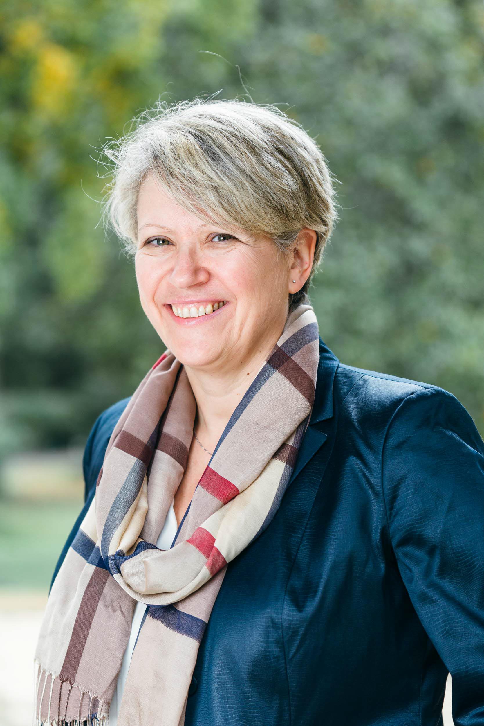 Une femme souriante aux cheveux courts et gris porte un blazer bleu foncé et une écharpe à carreaux aux nuances de beige, de marron et de rouge. Elle se tient à l'extérieur avec un arrière-plan vert flou à Bordeaux, suggérant un parc ou un jardin. Le temps semble doux et l'atmosphère générale est agréable et naturelle pour ce portrait professionnel.