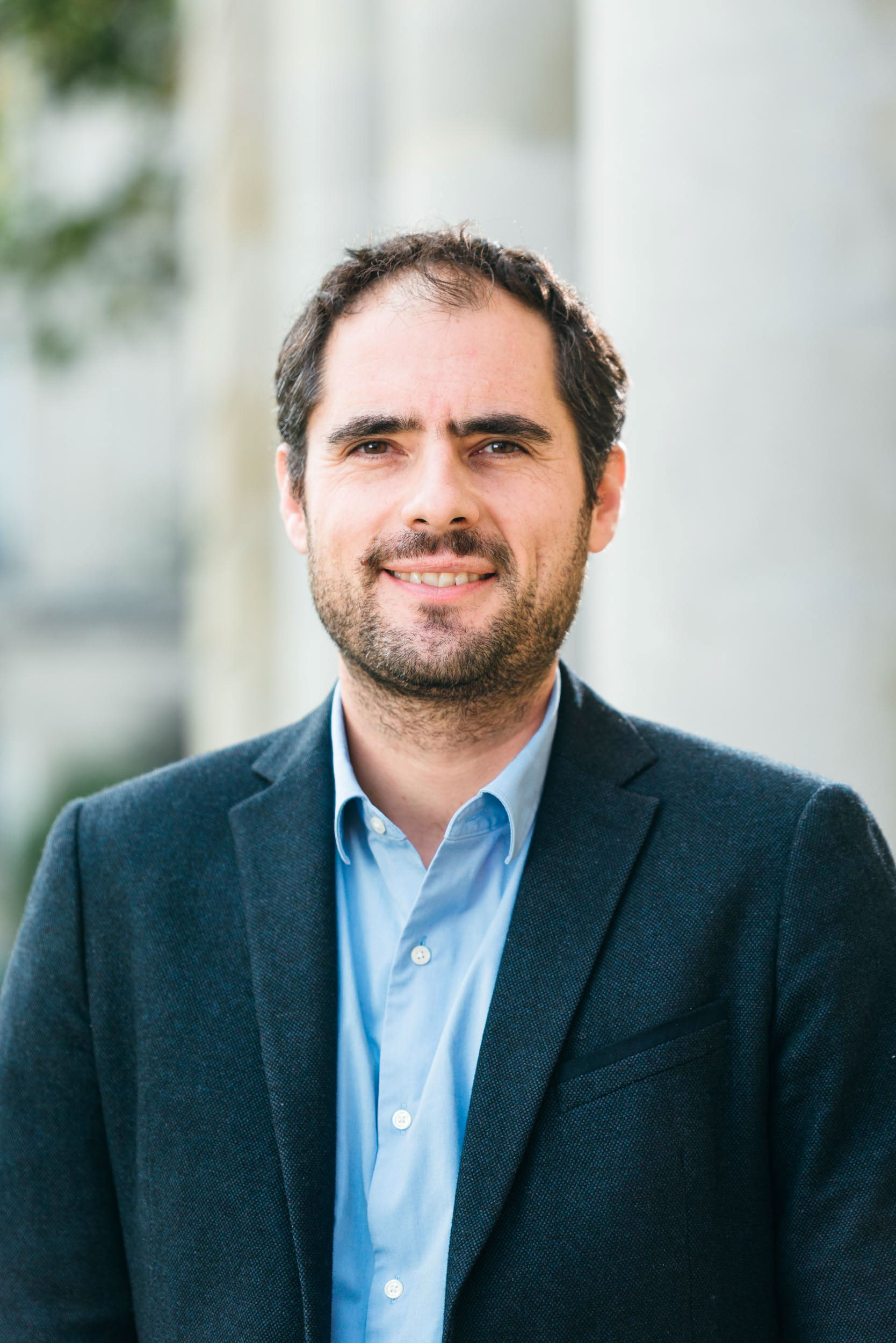 Un homme aux cheveux noirs courts et à la barbe hirsute se tient debout et sourit légèrement sur ce portrait d'entreprise. Il porte une chemise bleue sous un blazer foncé. L'arrière-plan est flou, montrant des colonnes de couleur claire et un peu de verdure, peut-être quelque part à Bordeaux ou en Gironde. L'éclairage est vif et semble naturel.